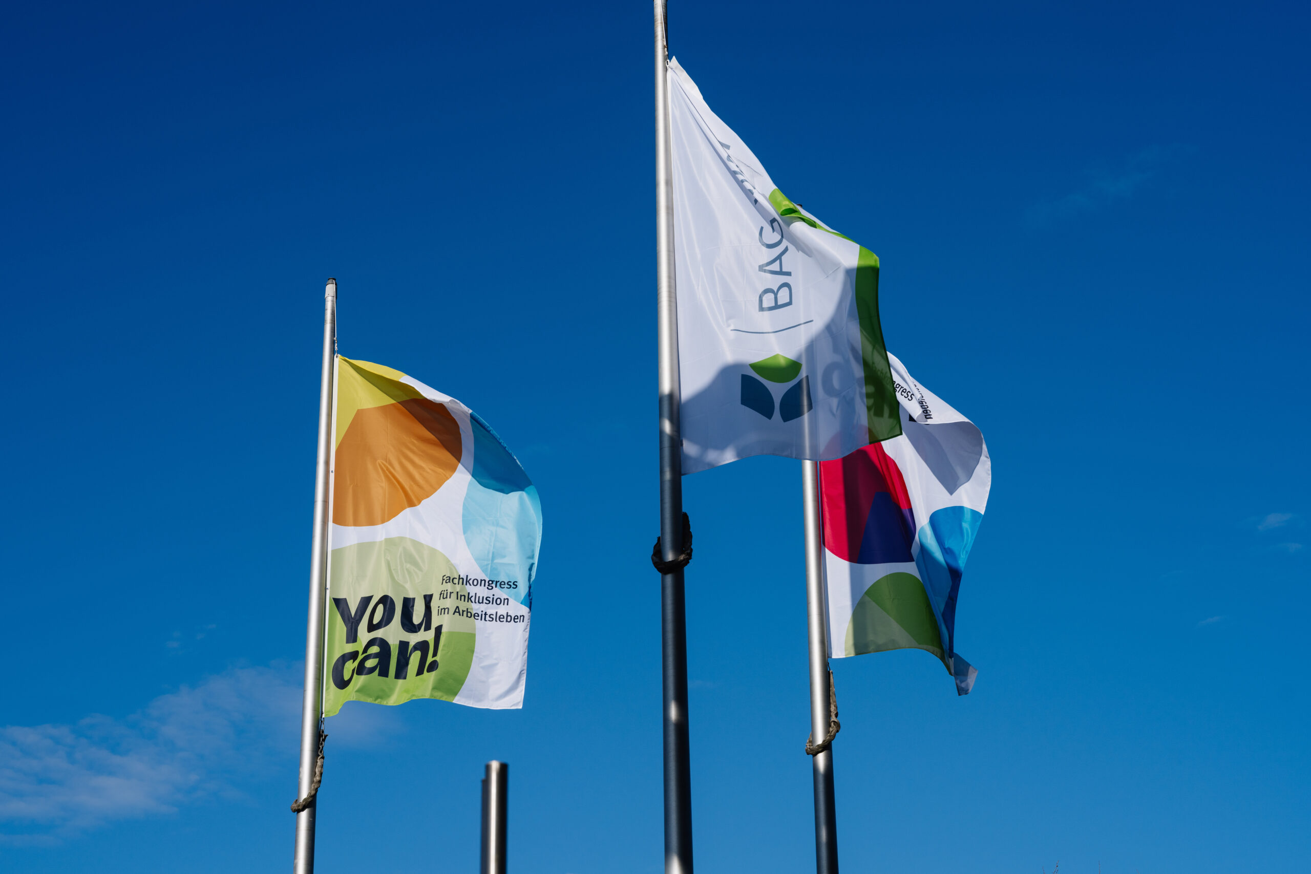 Das Foto zeigt drei Fahnen vor blauem Himmel. Die Fahnen tragen die Logos der BAG Wfbm und der Veranstaltung "You can! Fachkongress für Inklusion im Arbeitsleben".