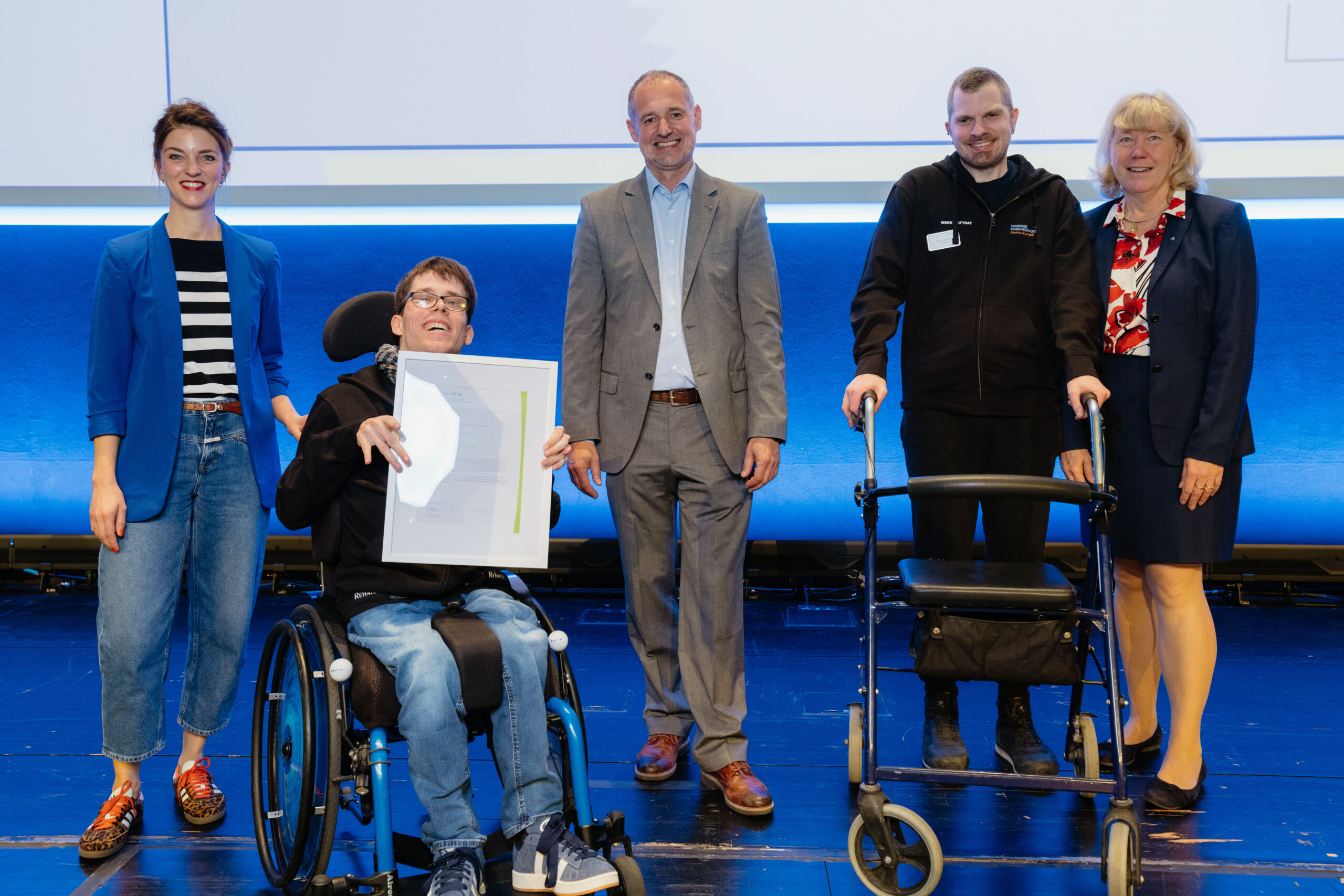 Das Foto zeigt fünf Personen auf einer Bühne. Sie sind nebeneinander und lächeln in die Kamera. Eine Person sitzt in einem Rollstuhl. Sie hält einen großen Bilderrahmen mit einer Urkunde in den Händen.