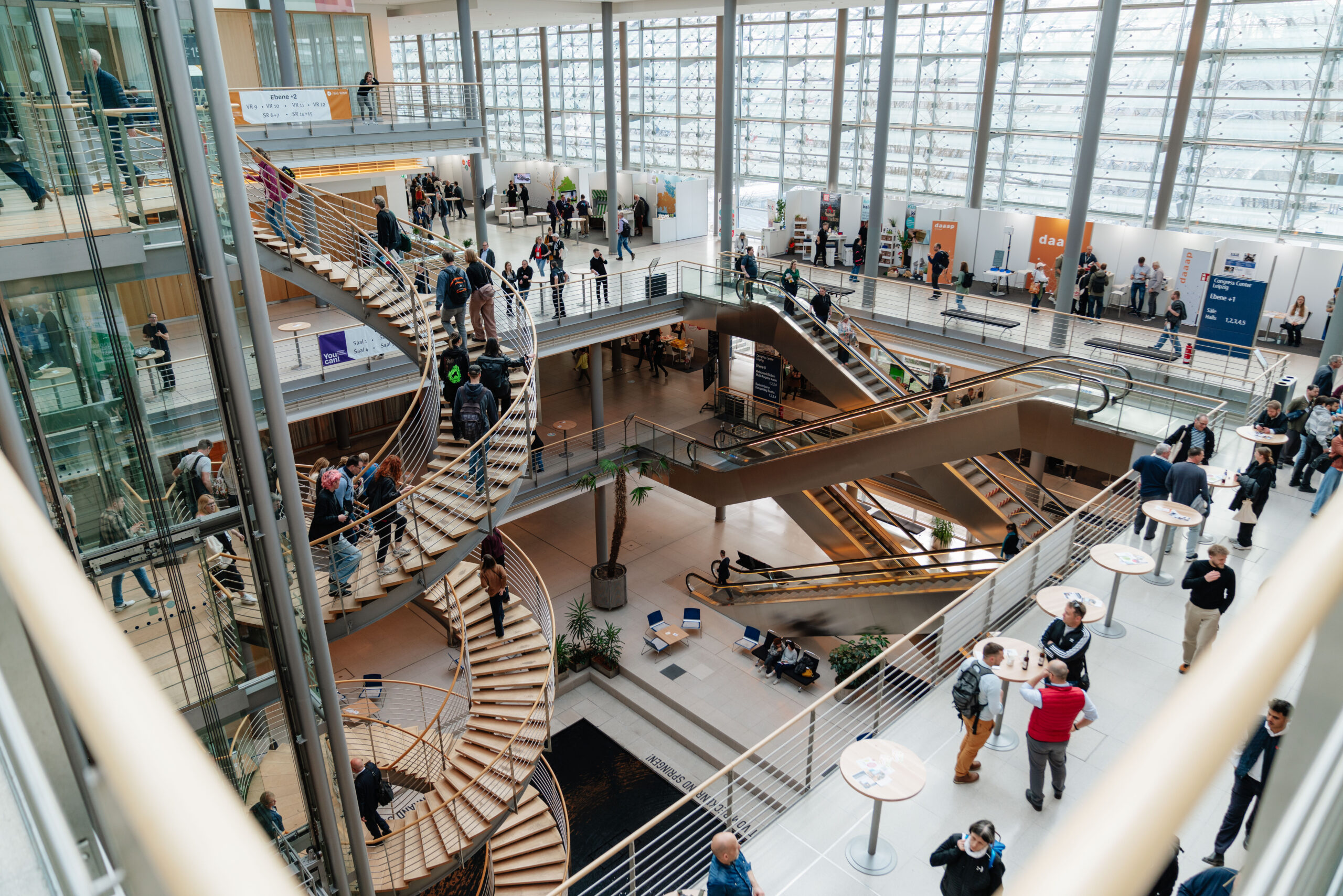 Das Foto zeigt ein offenes Treppenhaus mit Wendeltreppen, Rolltreppen und Fahrstühlen. Auf den einzelnen Etagen sind Menschen und Messestände erkennbar. Im Hintergrund ist eine große Glasfront.