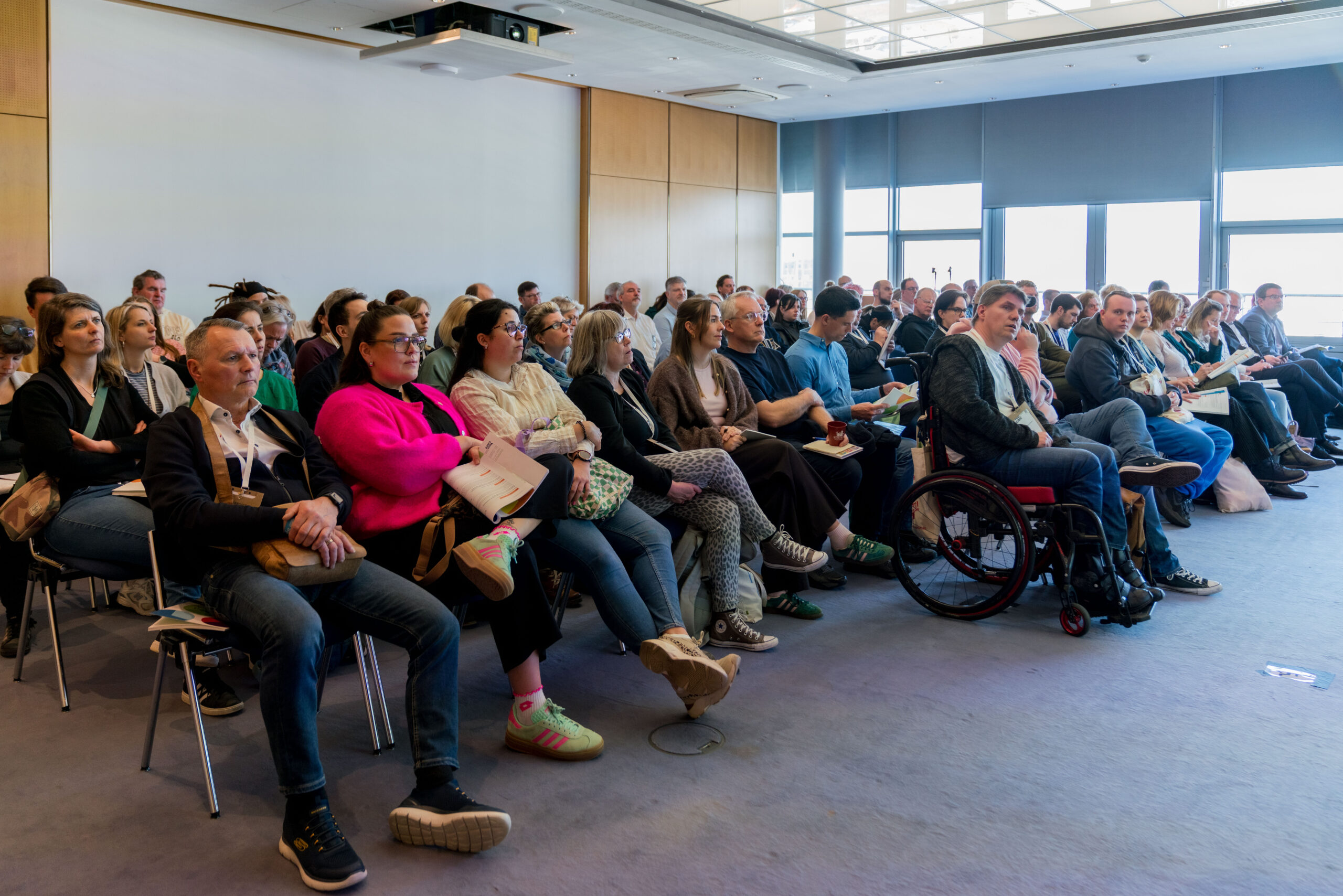 Das Foto zeigt eine große Zahl von Menschen, die in einem Raum in Stuhlreihen sitzen. Eine Person nutzt einen Rollstuhl, Alle blicken konzentriert geradeaus.