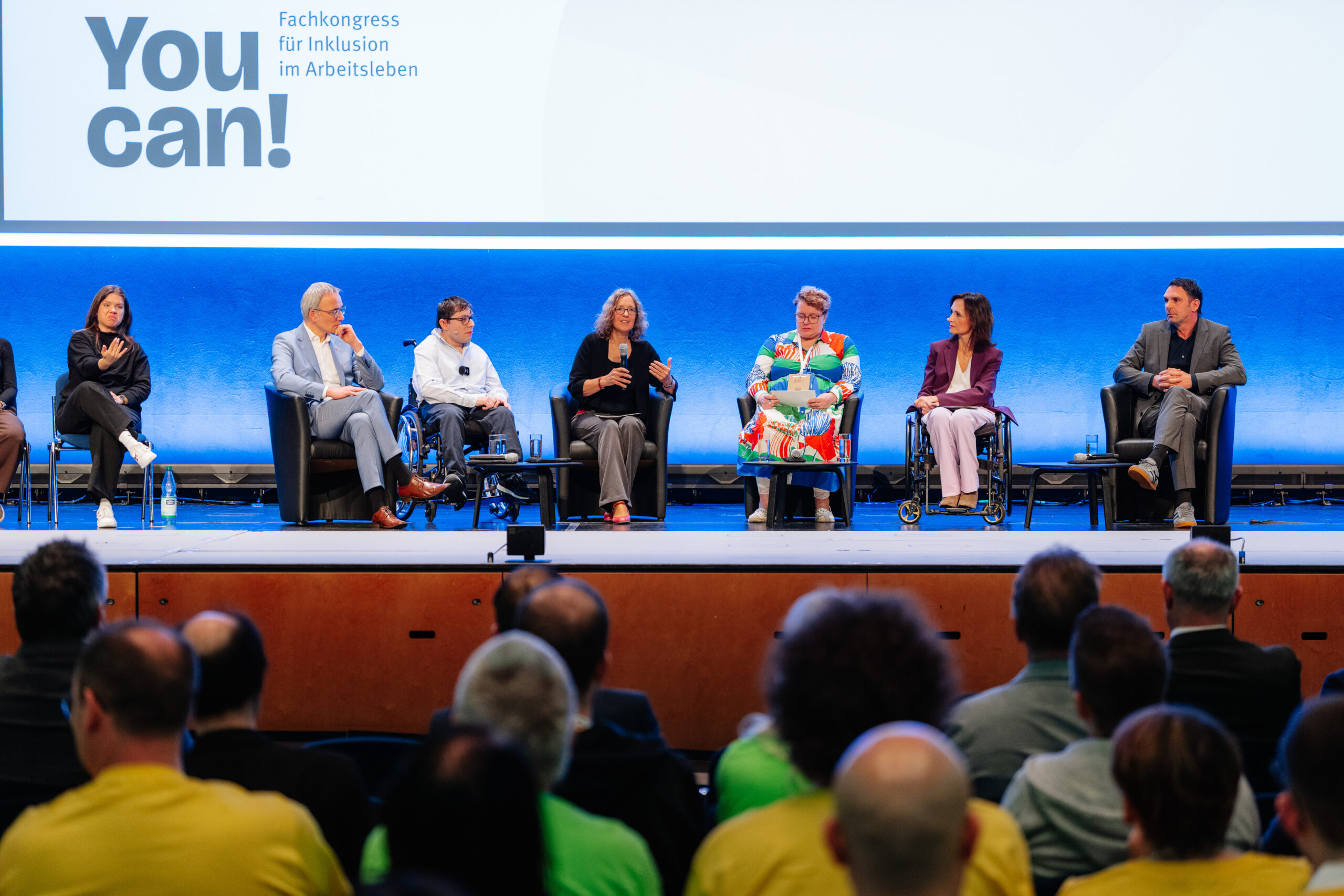 Das Foto zeigt mehrere Personen auf einer Bühne sitzend. Zwei Personen nutzen einen Rollstuhl. Ganz links ist eine Person, die Gebärdensprache verwendet. In der Mitte ist eine Person, die in ein Mikrofon spricht. Die übrigen Personen schauen sie an oder schauen in Richtuing des Publikums, das vor der Bühne in Reihen sitzt. Im Hintergrund ist eine Leinwand mit dem Logo der Veranstaltung You can!