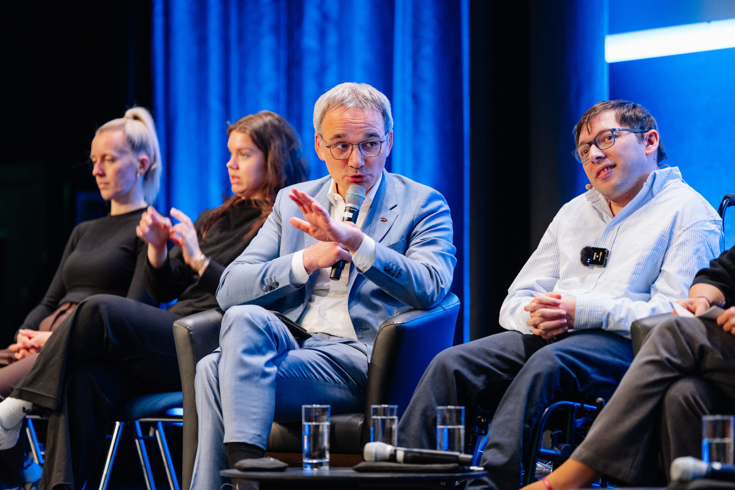 Das Foto zeigt eine Podiumsdiskussion. Im Fokus der Aufnahme sind ein Mann in hellblauem Anzug und mit Brille, der in einem Sessel sitzt, sowie ein Mann mit Brille und weißem Hemd, der in einem Rollstuhl sitzt. Der Mann im blauen Anzug spricht in ein Mikrofon und hat seine linke Hand leicht angehoben. Im Hintergrund ist eine Frau, die in Gebärdensprache dolmetscht.