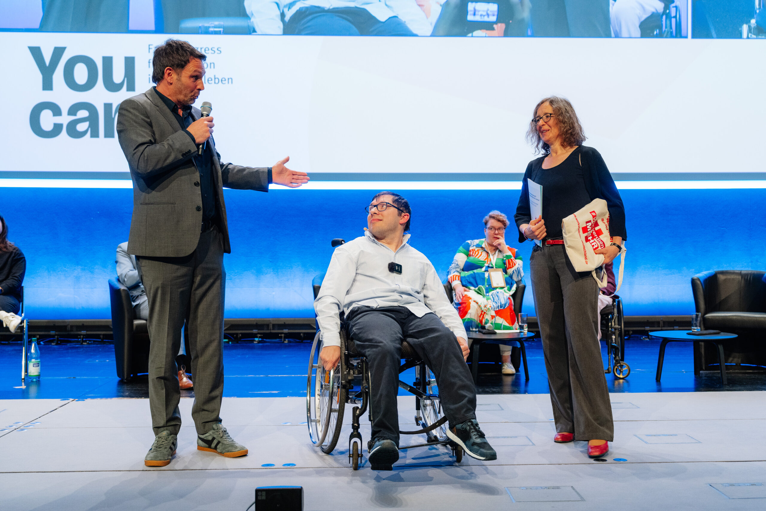 Das Foto zeigt drei Personen auf einer Bühne. Rechts ist ein Mann, der in ein Mirkofon spricht und sich mit leicht erhobener linker Hand in Richtung der anderen beiden Personen wendet. Der Mann in der Mitte nutzt einen Rollstuhl und blickt den Mann links lächelnd an. Rechts steht eine Frau. Auch sie lächelt. Unter den Armen hält sie einen gefüllten Jutebeutel mit der Aufschrift "Wir verdienen mehr" und eine Papier-Mappe.