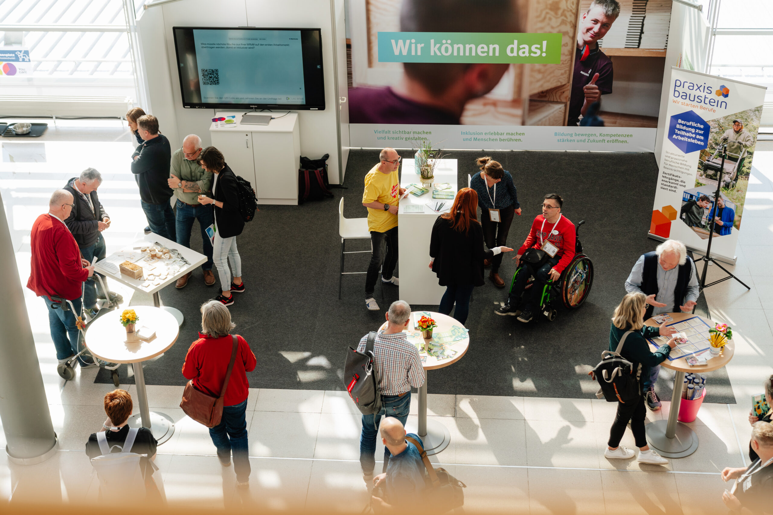 Das Foto zeigt Menschen an einem Messestand, die von oben aufgenommen sind. Sie stehen in kleinen Gruppen zusammen und unterhalten sich.
