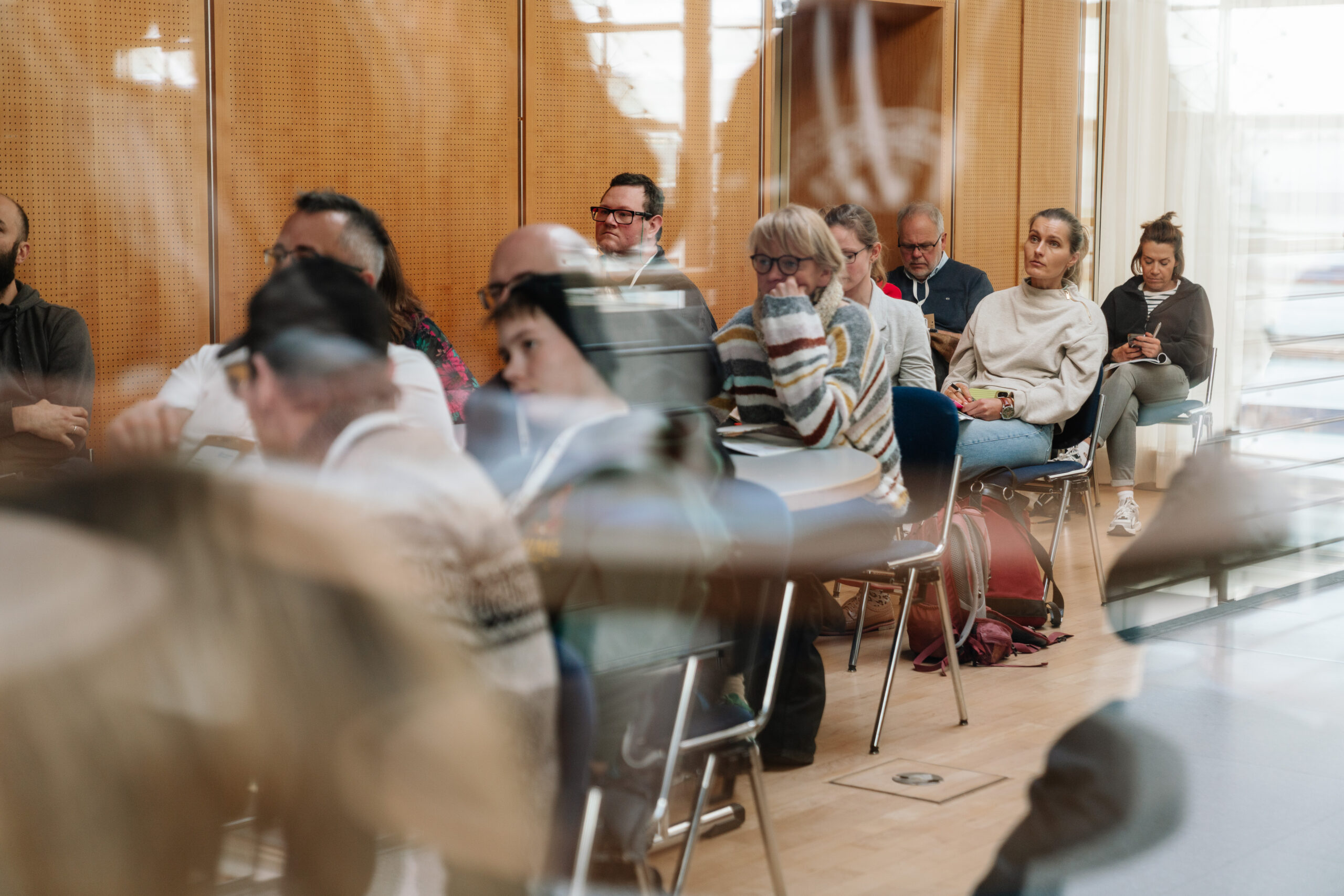 Das Foto zeigt hinter einer Glasscheibe mehrere Personen, die in Stuhlreihen sitzen. Sie blicken konzentriert nach vorne.