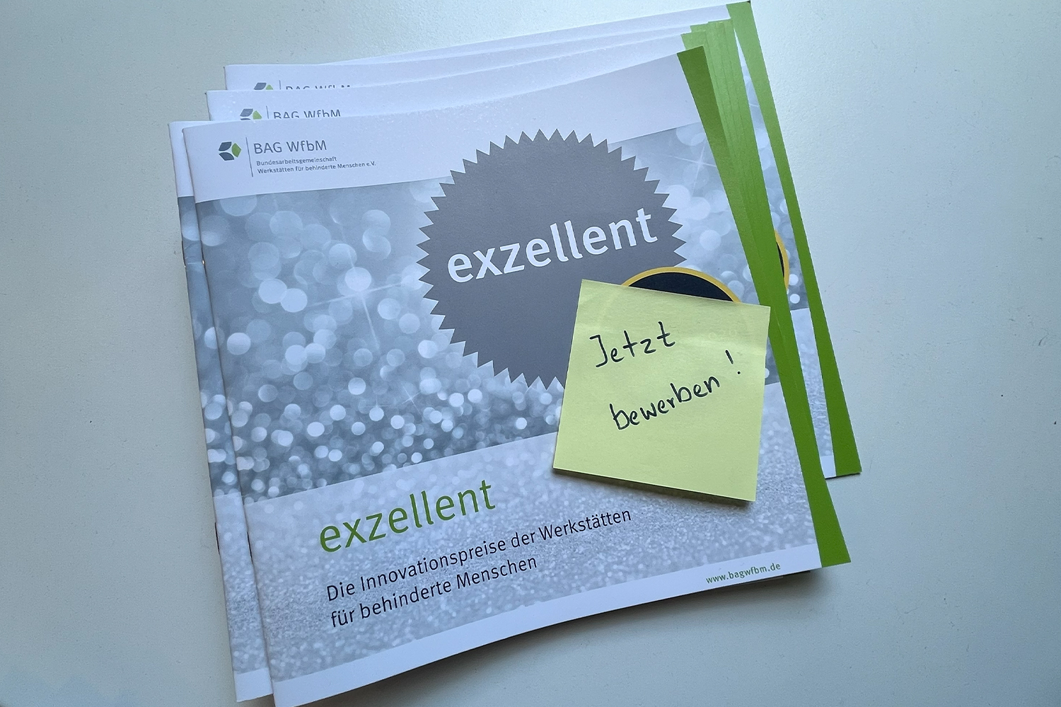 Das Foto zeigt mehrere Broschüren der exzellent-Preise der BAG WfbM. Die Broschüren liegen übereinander gestapelt auf einem Tisch. In der rechten unteren Ecke der obersten Broschüre klebt ein quadartisches Post-It mit der Aufschrift Jetzt bewerben!