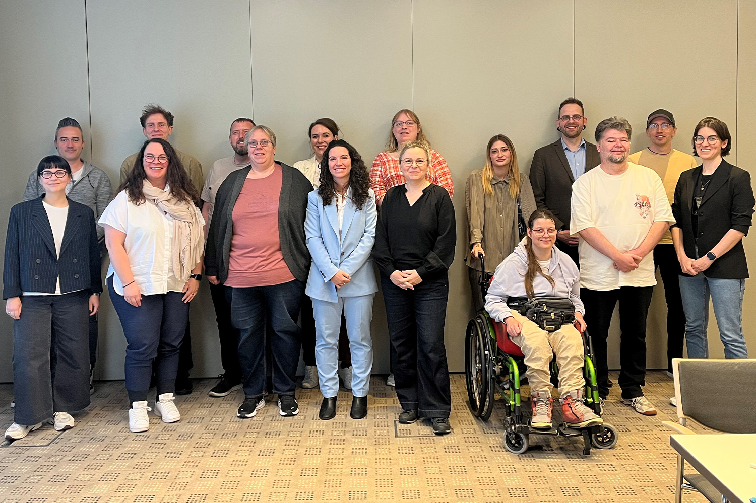 Das Foto zeigt eine Gruppe von 16 Personen. Sie stehen in zwei Reihen hintereinander vor einer Wand in einem Konferenzraum. Eine Person nutzt einen Rollstuhl. Alle lächeln nach vorne in die Kamera.