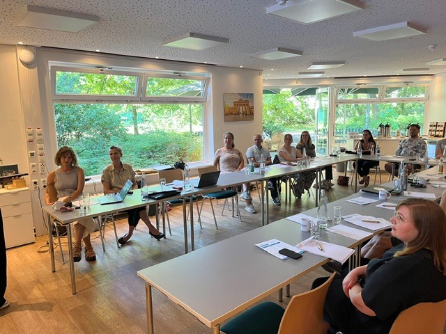 Das Foto zeigt einen Seminarraum, in dem viele Menschen an Tischen sitzen. Die Tische sind in U-Form ausgerichtet, sodass alle Menschen nach vorne blicken können. Auf den Tischen stehen Getränke und Laptops und es liegen Papiere darauf.