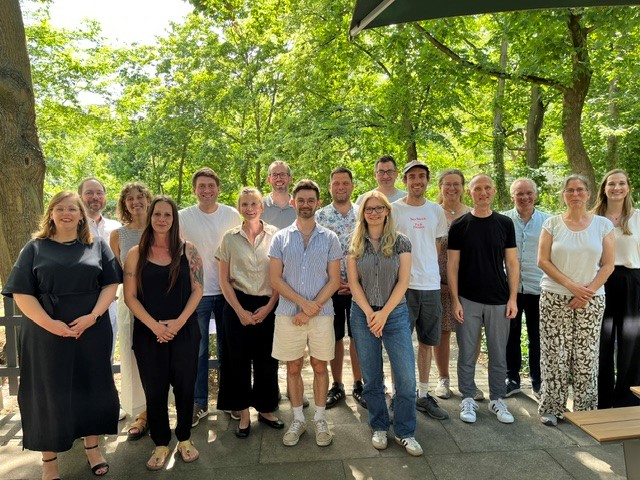 Das Foto zeigt eine Gruppe von Menschen im Freien. Sie stehen in mehreren Reihen neben- und hintereinander. Alle lachen geradeaus in die Kamera. Im Hintergrund sind Bäume mit dichtem Laub.