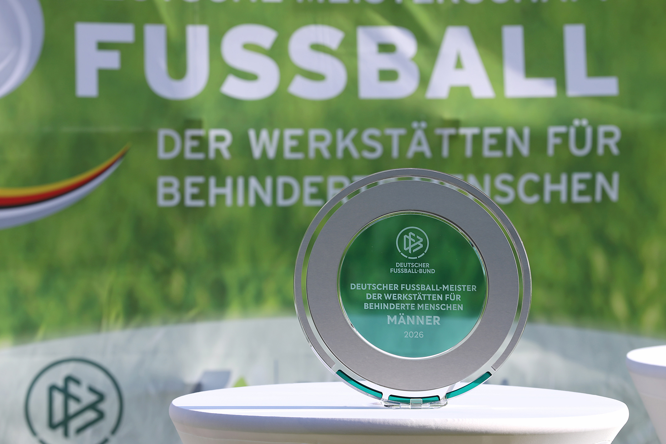Das Foto zeigt die Meisterplakette zur Deutschen Fußball-Meisterschaft der Werkstätten 2026. Sie steht auf einem Tisch mit weißer Tischdecke. Im Hintergrund ist ein Werbebanner des Turniers.
