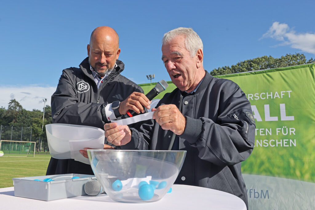 Das Foto zeigt zwei Männer an einem Stehtisch im Freien. Der Mann links im Bild hält ein weißen Blatt Papier in der einen und ein Mikrofon in der anderen Hand. Der Mann rechts spricht in das Mikrofon, während er Zettel aus Kugeln zieht, die sich in einer Schale vor ihm befinden.