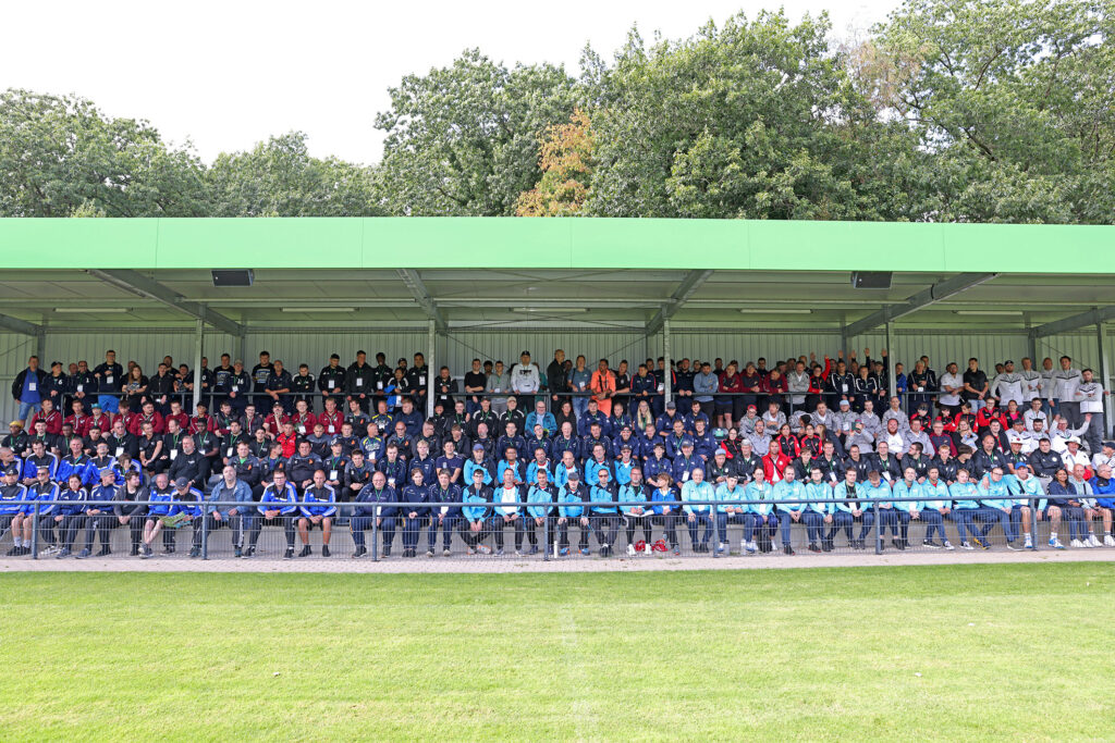 Das Foto zeigt eine langgezogene Tribüne neben einem Fußballplatz. Auf der Tribüne haben viele Menschen Platz genommen. Sie tragen bunte Fußball-Outfits.
