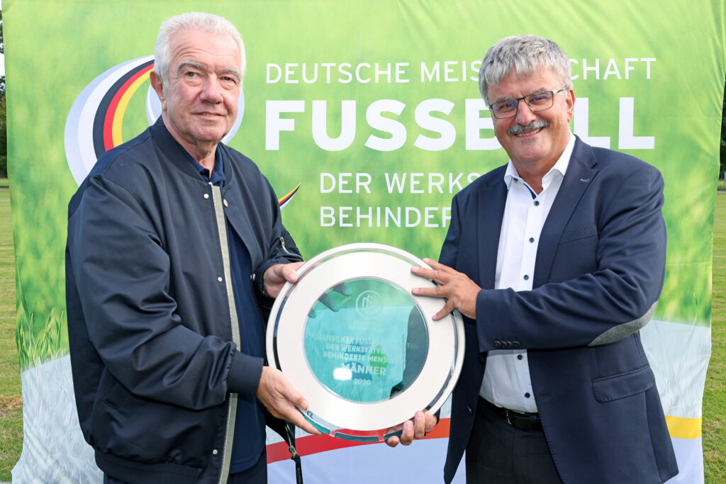Das Foto zeigt zwei Männer vor einem Werbebanner der Deutschen Fußball-Meisterschaft der Werkstätten. Die Männer halten gemeinsam eine Meisterschale in den Händen. Beide lachen geradeaus in die Kamera.