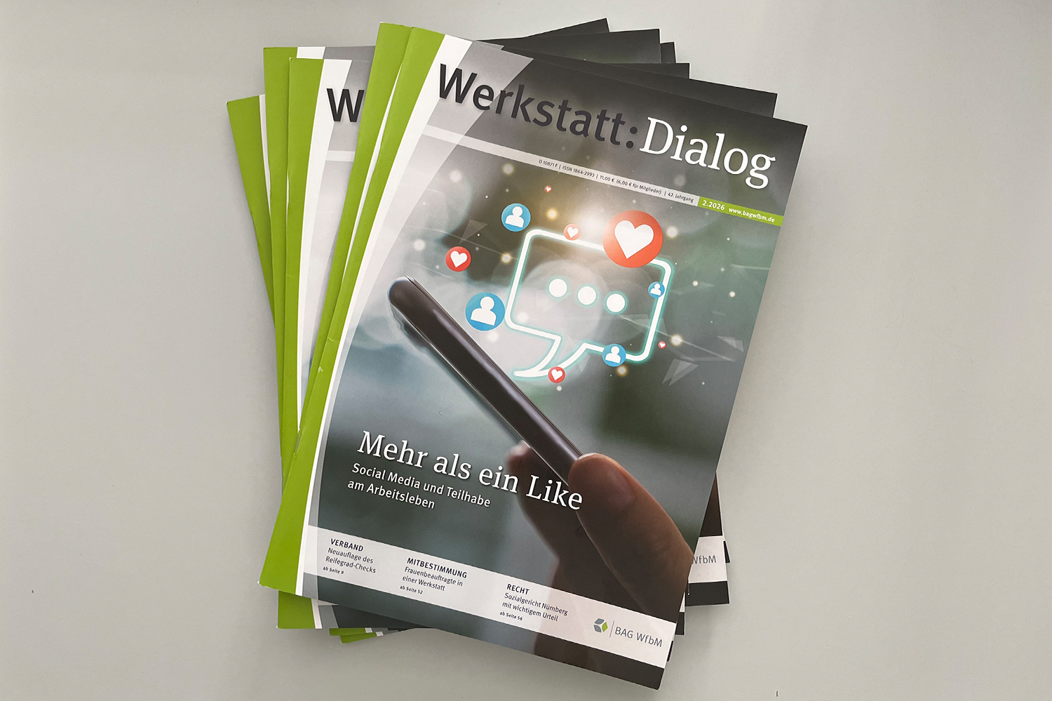 Das Foto zeigt einen Stapel mit Magazinen auf einer hellgrauen Flächen. Bei den Magazinen handelt es sich um die Ausgabe 2.2026 des Werkstatt:Dialogs der BAG WfbM. Die Magazine liegen so übereinander, dass jeweils die oberen linken Ecken erkennbar sind. Bei dem obersten Magazin ist das Cover des Heftes vollständig sichtbar. Unten links steht der Titel "Mehr als ein Like Social Media und Teilhabe am Arbeitsleben". Im Hintergrund ist ein Smartphone sichtbar, das von einer menschlichen Hand gehalten wird. Über dem Smartphone erscheinen mehrere Icons aus dem Bereich Social Media, darunter kleine Herzen und eine größere Sprechblase. Es sieht aus, als würden die Icons aus dem Smartphone aufsteigen.