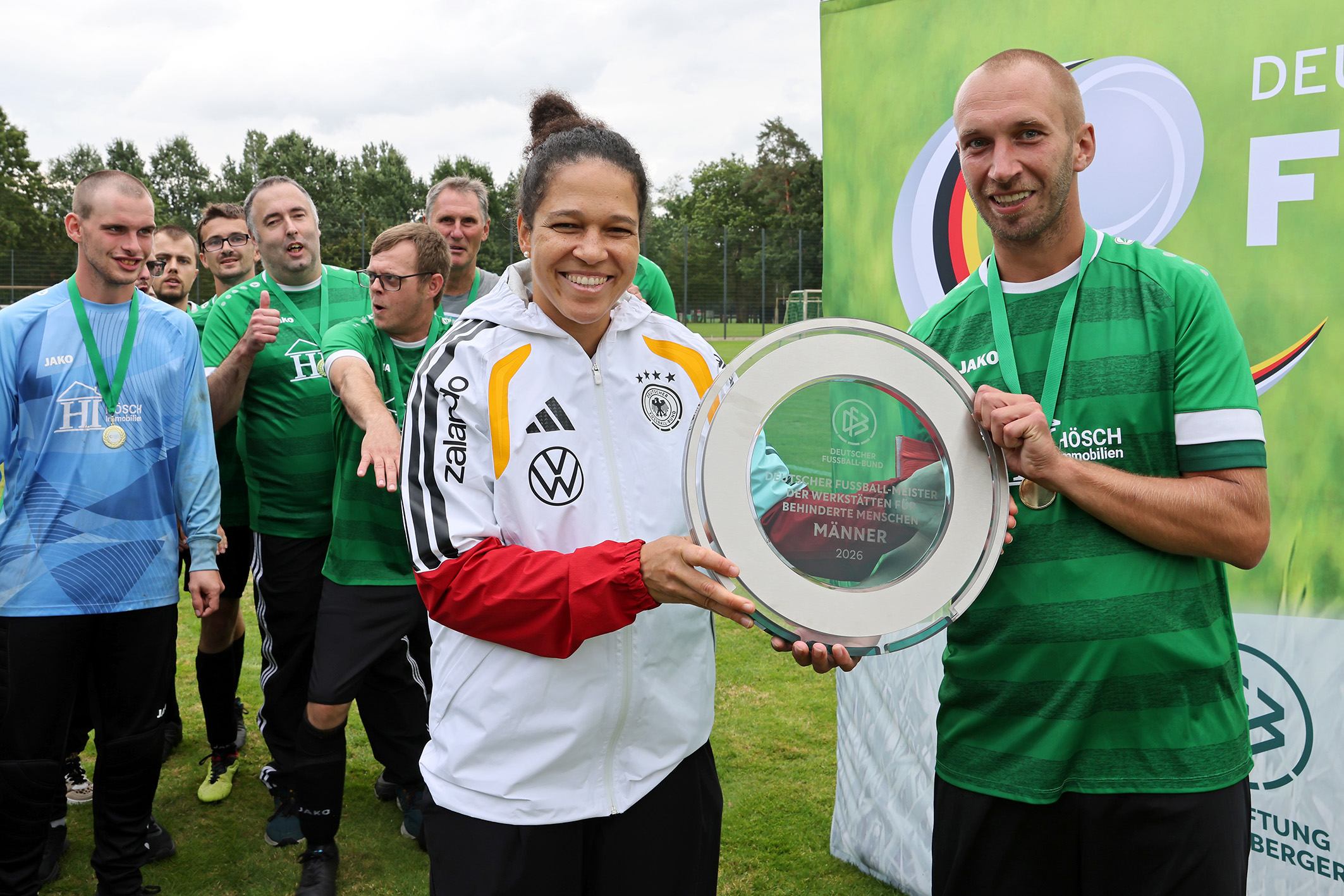 Das Foto zeigt die ehemalige Fußball-Nationalspielerin Celia Šašić bei der Übergabe der DFB-Meisterplakette an einen Spieler der Wefa Ahonr. Im Hintergrund sind weitere Mitglieder des Teams, die Medaillen um den Hals tragen. Sie wirken ausgelassen.