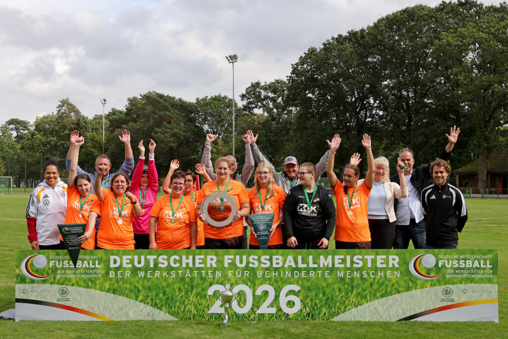Das Foto zeigt die Spielerinnen der Landesauswahl Rheinland-Pfalz mit Trainern und Unterstützern. Sie stehen hinter einem Banner mit der Aufschrift "Deutscher Fußballmeister der Werkstätten für behinderte Menschen 2026". Alle Menschen auf dem Foto lachen, jublen und schauen in die Kamera.