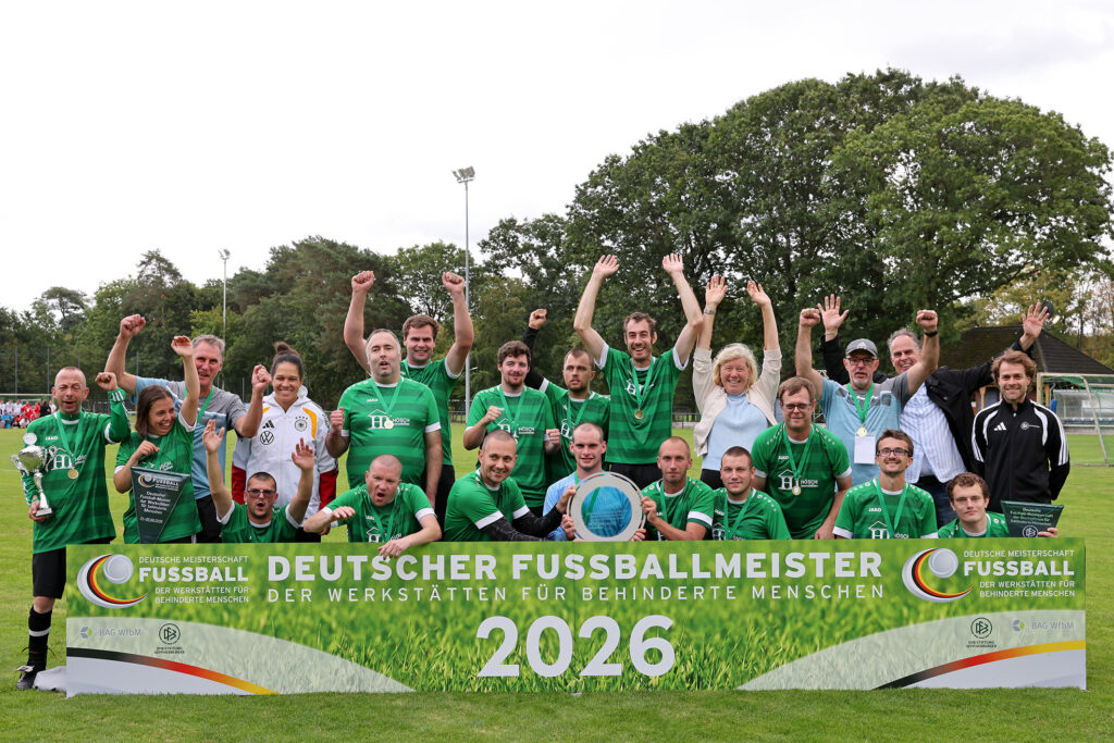 Das Foto zeigt die Spieler der Wefa Ahorn mit Trainern und Unterstützern. Sie stehen hinter einem Banner mit der Aufschrift "Deutscher Fußballmeister der Werkstätten für behinderte Menschen 2026". Alle Menschen auf dem Foto lachen, jublen und schauen in die Kamera. In der Mitte sind Spieler, die eine Meisterplakette in den Händen halten.