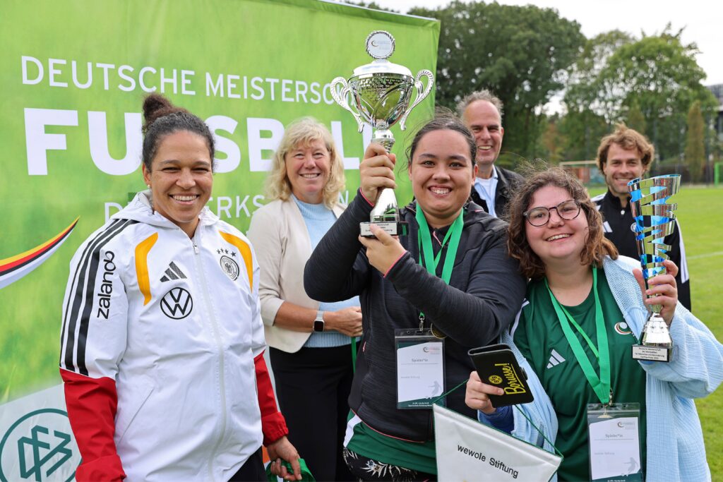 Das Foto zeigt zwei Fußballspielerinnen, die beide einen Pokal in der Hand halten und in die Kamera lachen. Um sie herum stehen die ehemalige Fußball-Nationalspielerin Celia Šašić, Andrea Stratmann von der BAG WfbM sowie Vertreter der DFB-Stiftung Sepp Herberger. Auch sie lachen in die Kamera.