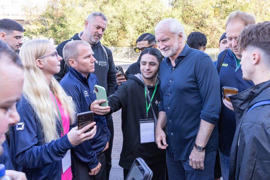 Das Foto zeigt Rudi Völler, umringt von Fußball-Spielern. Sie machen Selfies mit ihm.