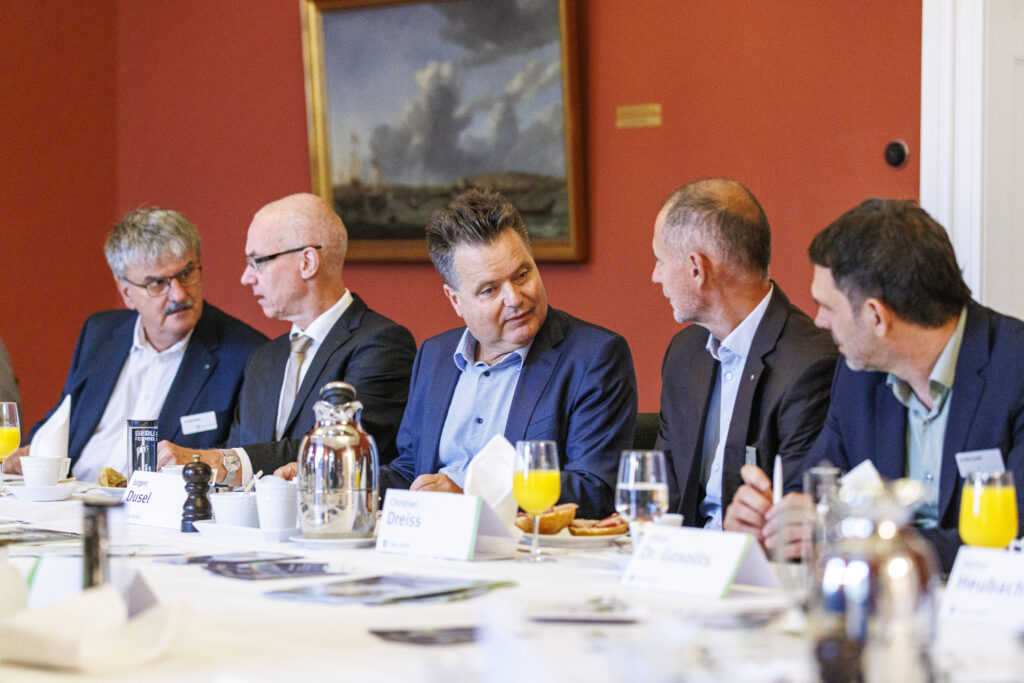 Das Foto zeigt fünf Männer, die in einer Reihe nebeneinander an einem Tisch sitzen. Alle tragen Anzüge und unterhalten sich angeregt miteinander. Vor ihnen auf dem Tisch steht Frühstücksgeschirr.