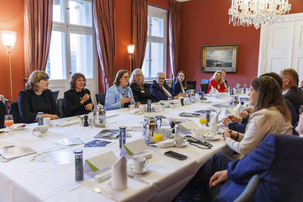 Das Foto zeigt mehrere Menschen, die zu beiden Seiten eines langen Tisches sitzen. Auf dem Tisch befinden sich Frühstücksgeschirr und Dokumente. Die Personen unterhalten sich angeregt miteinander.