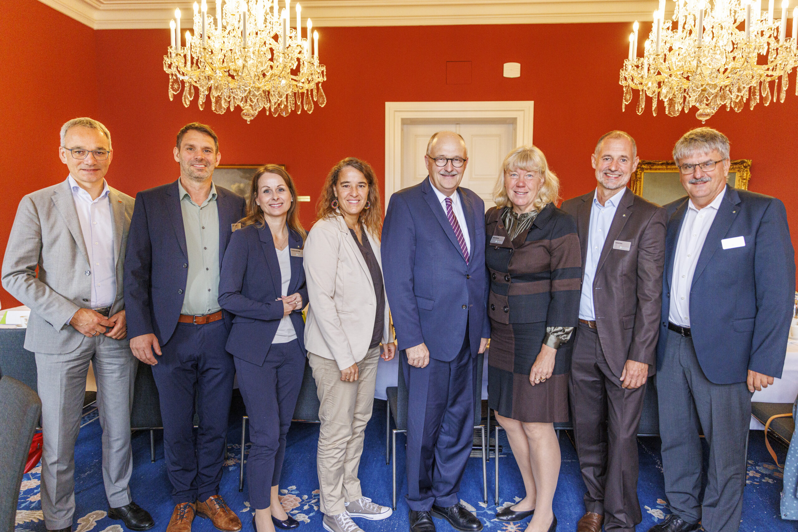 Das Foto ist ein Gruppenbild. Es zeigt acht Personen, die sich nebeneinander aufgestellt haben. Alle sind schick gekleidet und lächeln geradeaus in die Kamera.