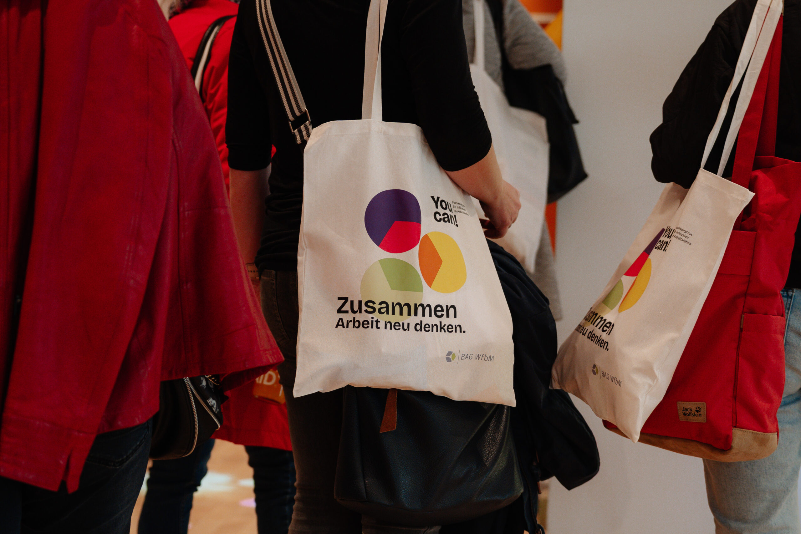 Das Foto zeigt eine Gruppe von Menschen. Ihre Köpfe und Beine sind nicht zu sehen. Mehrere von ihnen tragen einen Jutebeutel über der Schulter. Auf diesem ist das Logo vom You can! Fachkongress abgebildet.